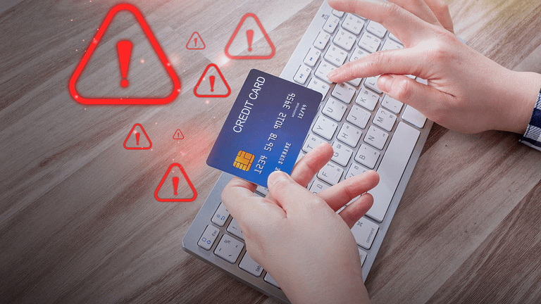 Image of hands using a keyboard with a credit card, surrounded by red warning symbols, indicating a potential fraud or security alert during an online transaction.