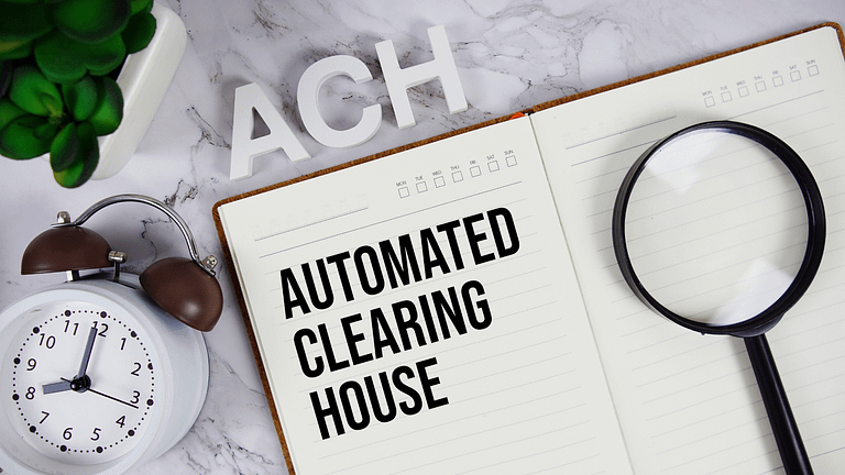 Image of an open notebook with "AUTOMATED CLEARING HOUSE" written, "ACH" blocks, a magnifying glass, clock showing 11:10, and a green plant on a marble surface.